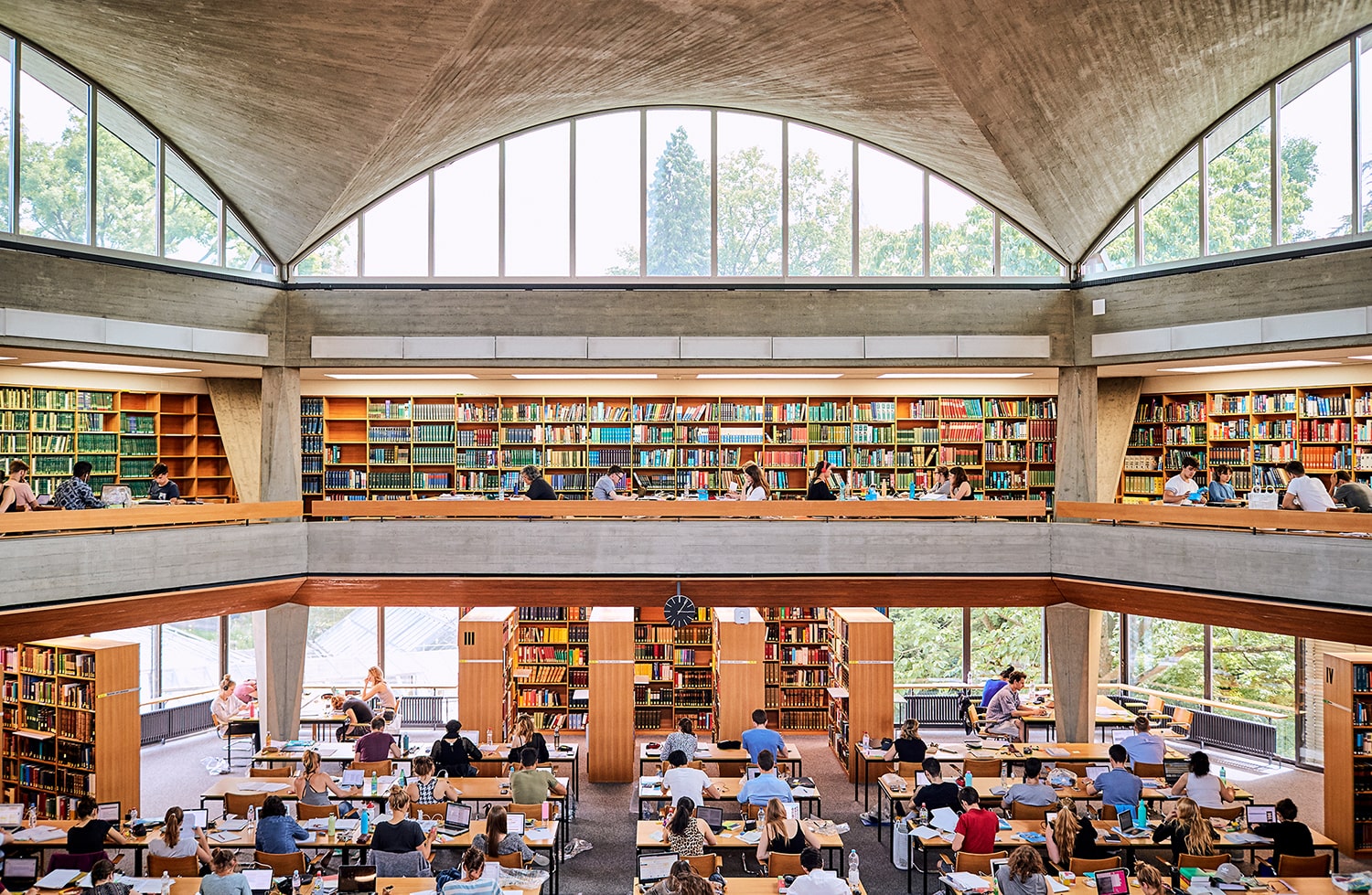 Viele Studierende lernen auf zwei Ebenen im grossen Lesesaal der Universitätsbibliothek. An den Wänden stehen Bücherregale und über den Lesesaal spannt sich eine geschwungene Beton-Decke mit grossen, halbrunden Fenstern.
